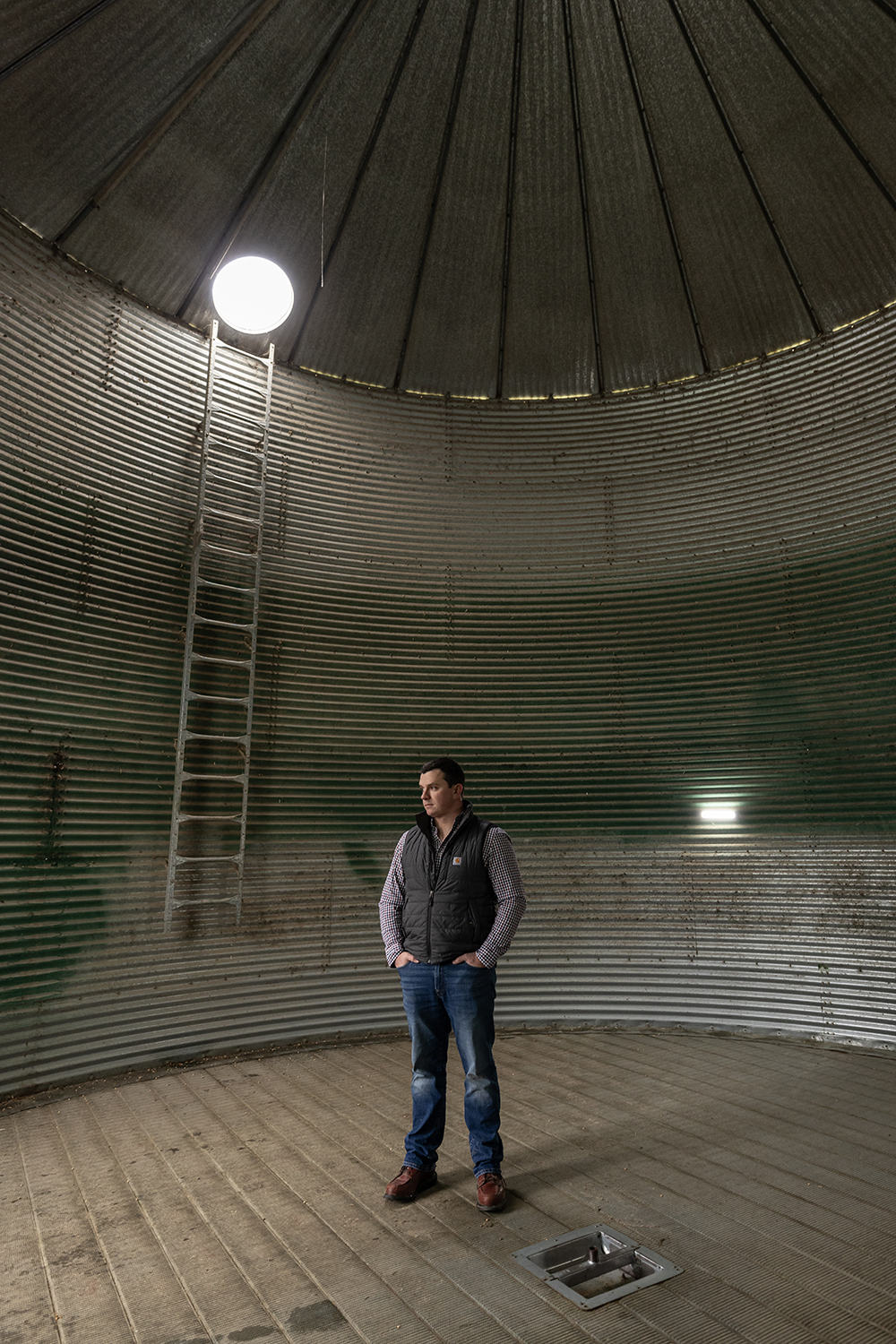 Will Hamman, a young white man wearing jeans and an insulated vest over his plaid button-down, stands inside an empty grain silo on his family farm in Ohio. The structure is corrugated metal maybe five times as wide as he is tall, with a peaked roof. A ladder and big flood light are too his right.