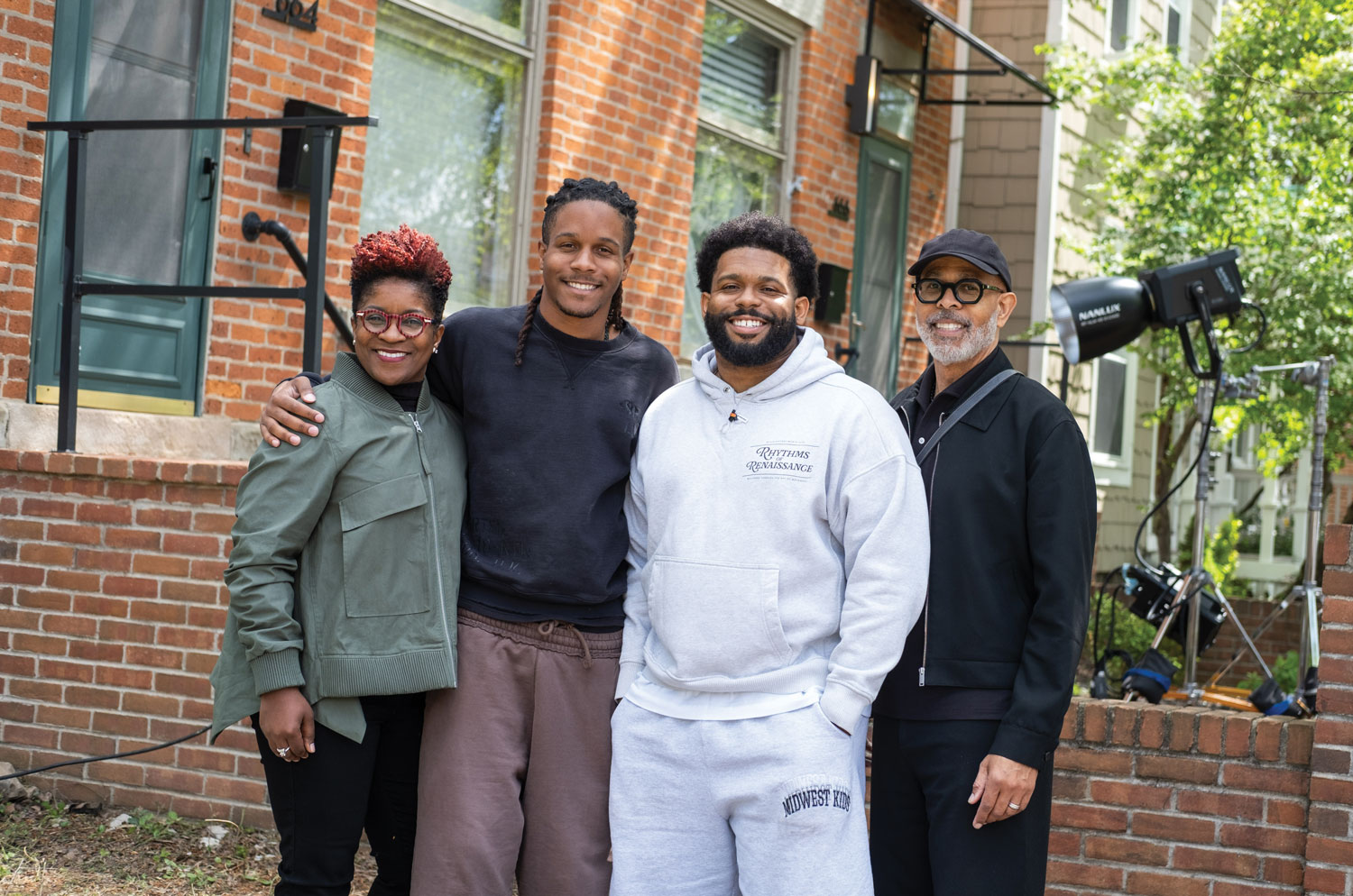 Justen poses for a photo with his family—his mom and dad and his brother. They all smile and stand close together, like they're not only comfortable together but enjoy each other. They stand in front of a brick double with lighting equipment behind them. 