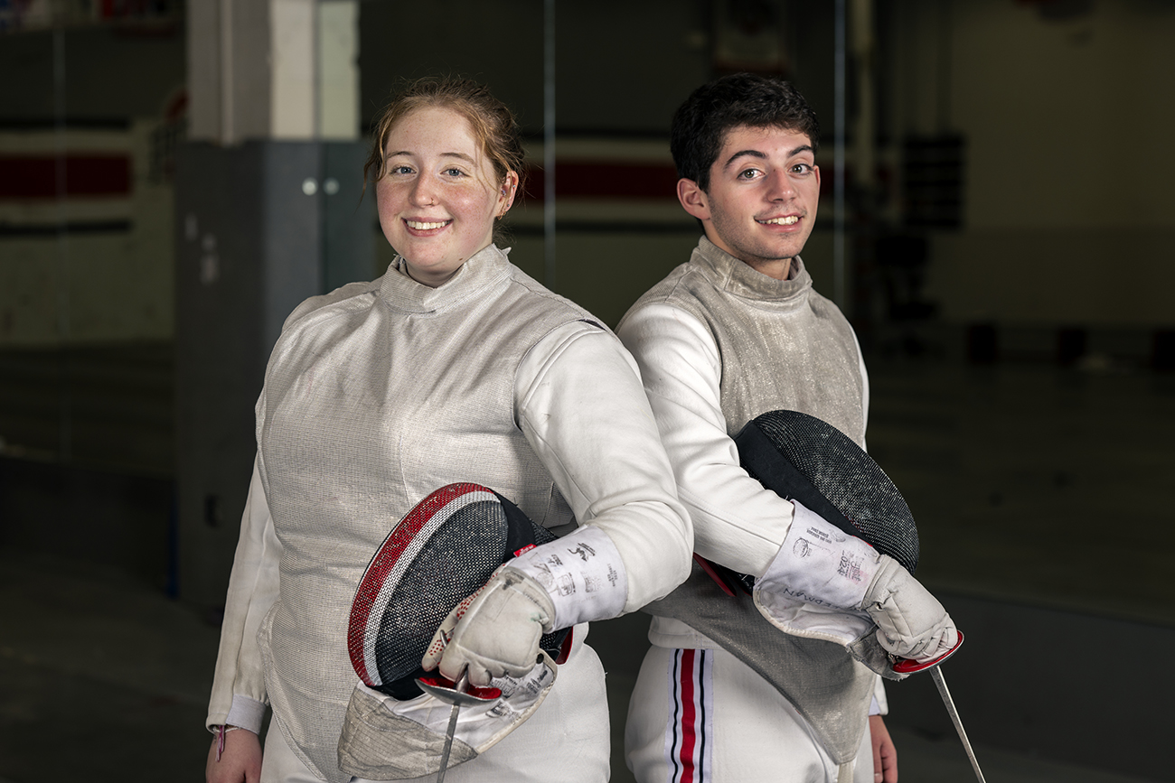 Siblings Miranda and Sammy Freedman smile as they pose for a photo in their fencing uniforms. Both are gray with use--they look like they've enjoyed a lot of competition time in them.