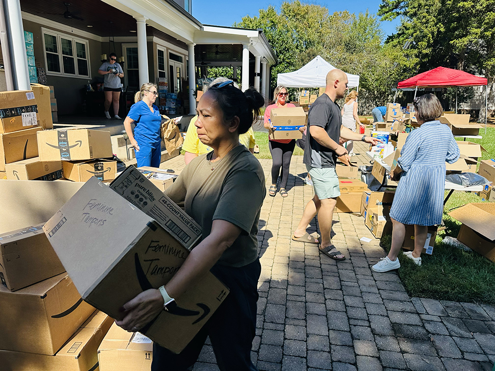 An assortment of people organize boxes in the back yard of a stately house. The sun shines brightly and people wear summer clothes