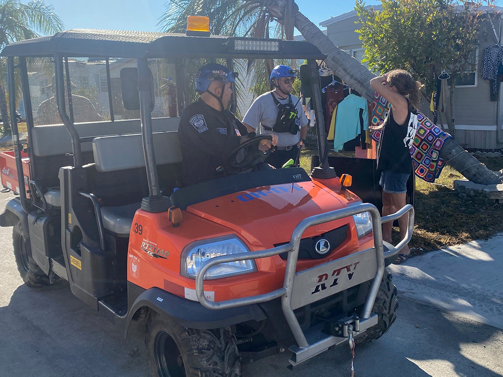 In front of a home, rescuers who came in on a suped-up four wheeler talk with a resident who experienced damage from a hurricane. The medical workers wear helmets and safety gear as the talk to the woman. In the background clothes and a blanket are drying on a fallen tree.