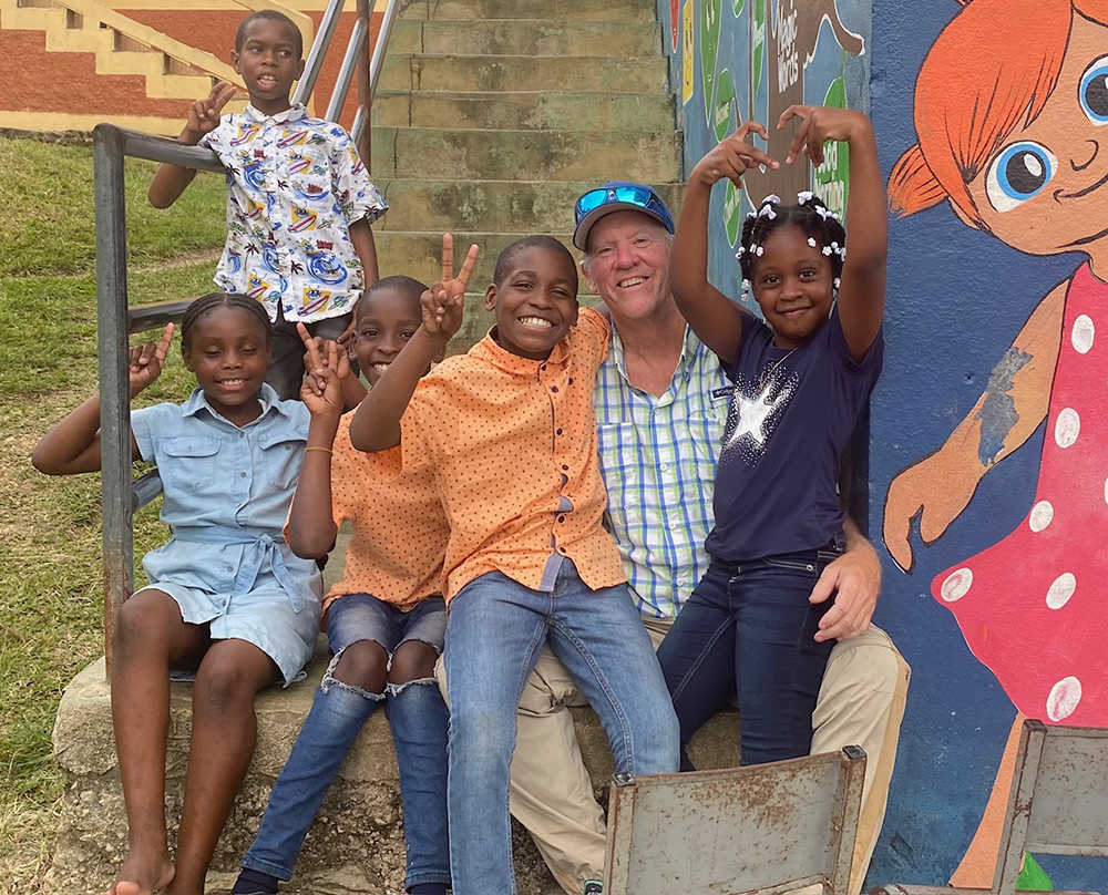 A man sits with five kids on stairs next to colorfully painted walls. They're all grinning and the kids are making peace or heart signs.