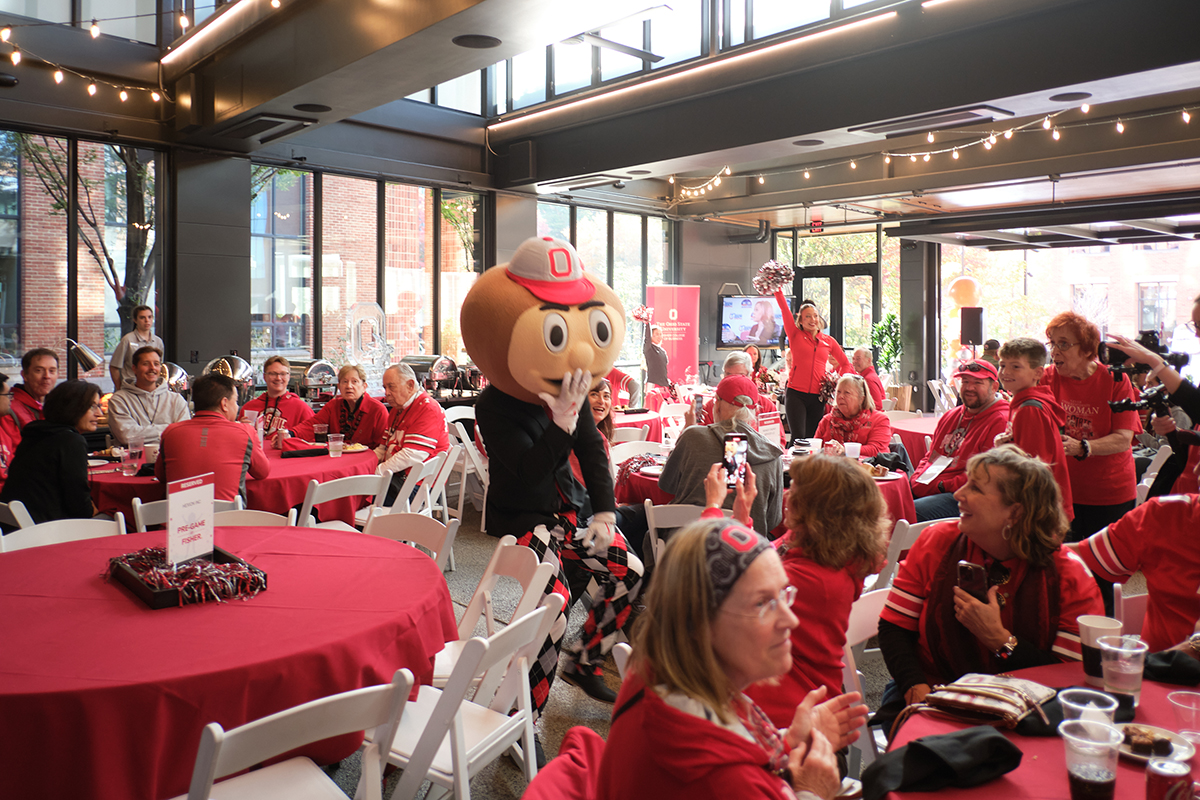 People clad in Ohio State gear sit at round tables inside a space wrapped in windows and filled with beautiful sunshine. They're all looking at Brutus Buckeye, whose hand is over his mouth as if laughing, and who wears a suitcoat and, for him, dressy pants with a diamond pattern. The crowd is laughing