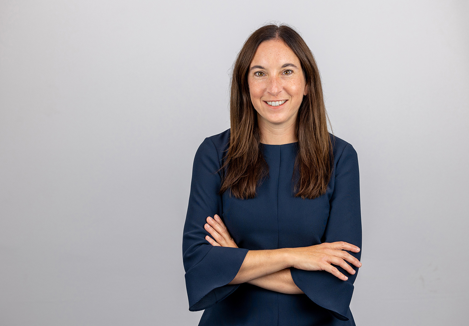 Margaret Kwoka, a friendly seeming white woman with freckles and long hair, poses for this photo with arms loosely crossed. She's standing in front of a plain background.