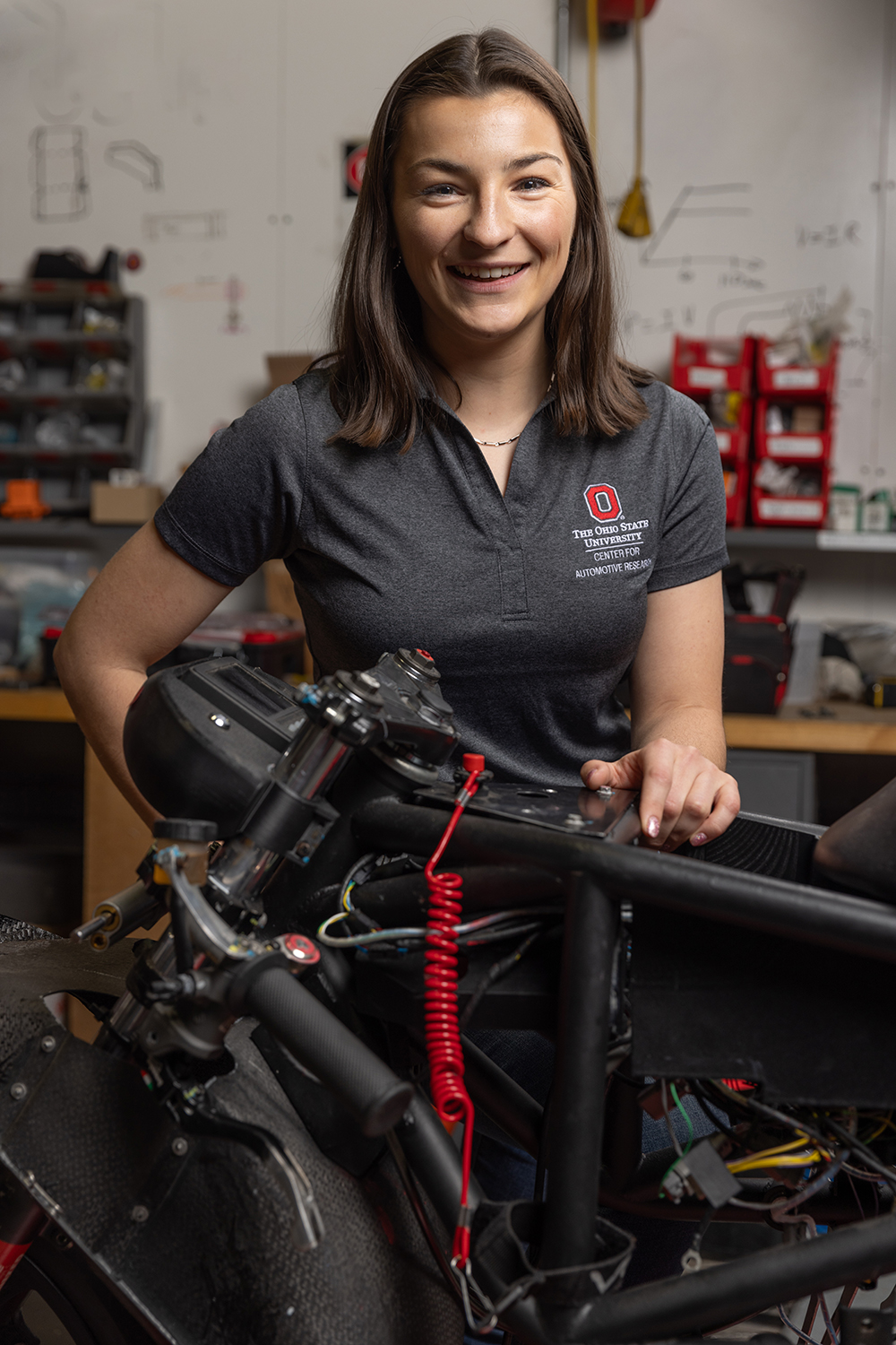 Laura Friedmann wears an Ohio State polo shirt branded Center for Automotive Research, as she smiles in this portrait in a motorcycle laboratory. She is a young woman with shoulder-length hair and her smile says she is genuinely thrilled to be photographed.