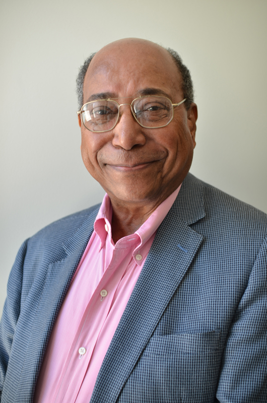 Noel Mayo smiles for a portrait, wearing glasses and a checked sportcoat, but no tie.