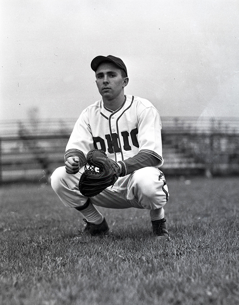 In a historical photo, a baseball player crouches and wears a serious expression as he is photographed. The front of his jersey says Ohio and his mitt is thick with an extra large thumb portion.