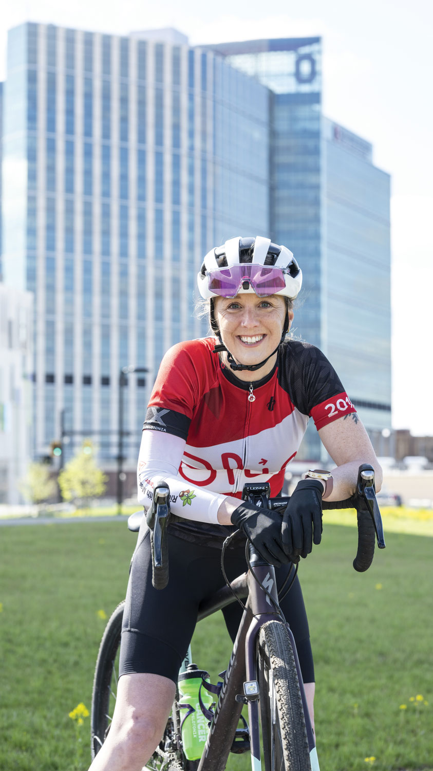 A white woman on her bicycle and wearing a riding helmet and jersey smiles as she poses for a photo in front of Ohio State's new hospital tower