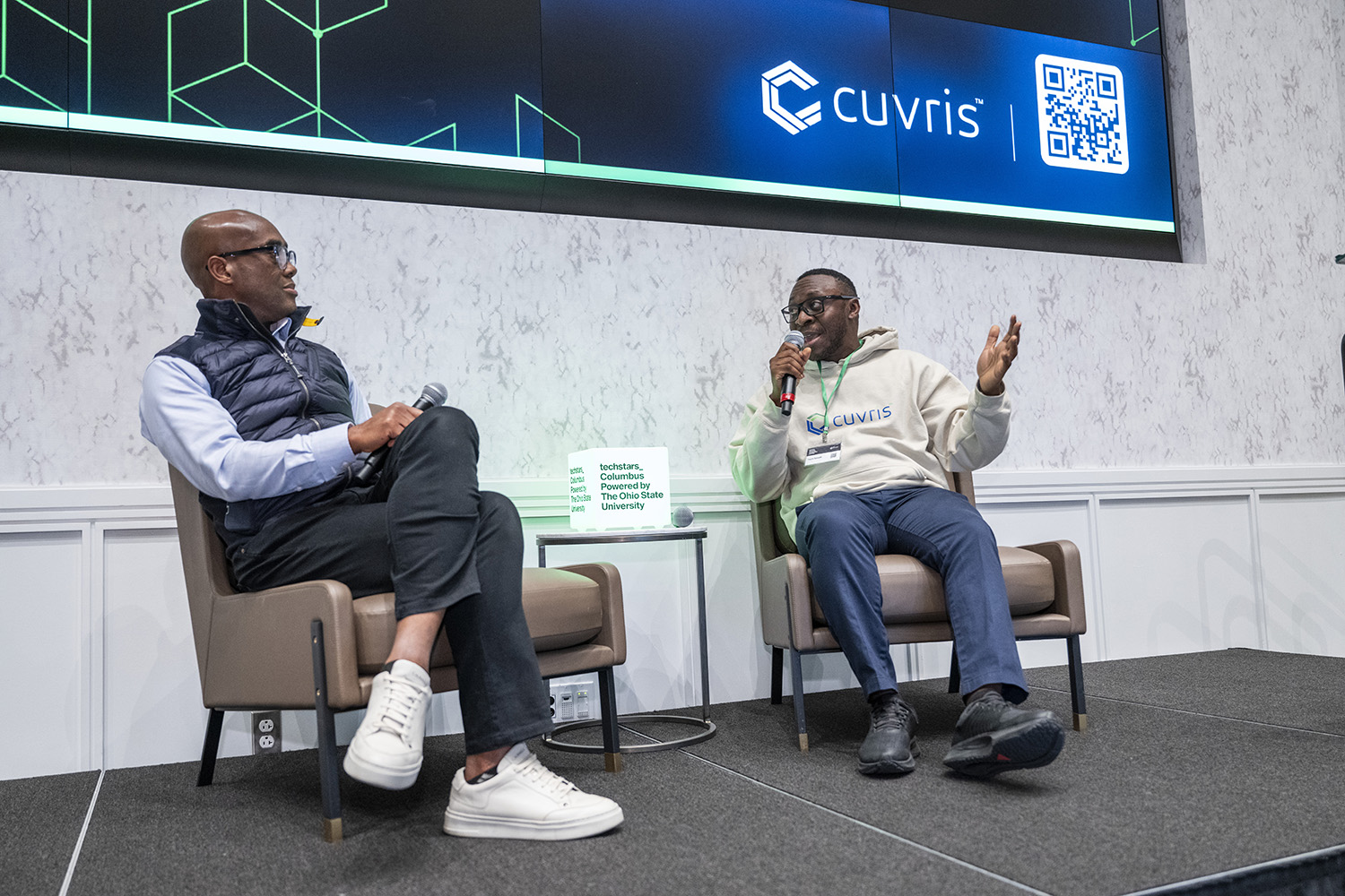 Two black men sit in chairs on a stage, having a discussion before an audience. Both hold microphones and wear similarly shaped and colored glasses. They make eye contact as Dami Sonoiki speaks.