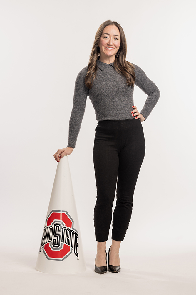 A pretty white woman with long hair and a friendly smile leans on a megaphone she used while a cheerleader in her Ohio State days. She's dressed in slim-fitting long-sleeved shirt and ankle-length pants.