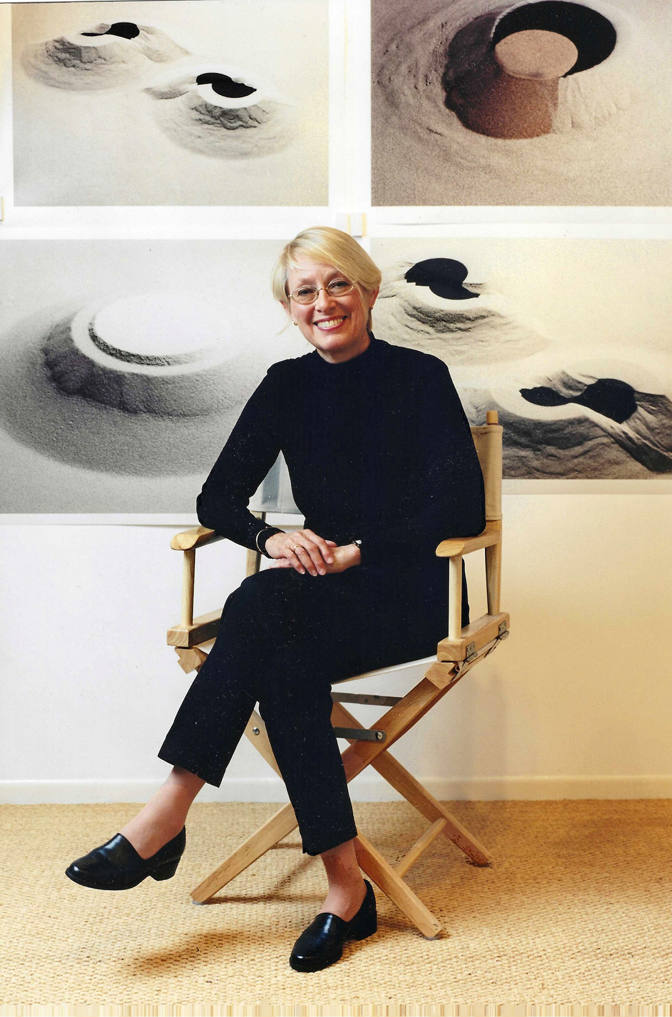 Connie Zehr, a white woman with short blond hair, oval glasses and a big smile, sits on a director's-style folding chair in front of photos of her works. They are all sand and they are mounded in different formations. They could be ancient or alien structures, or foot prints into mounded sand--they're abstract and so open to interpretation.