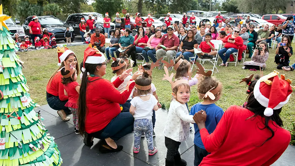 A group of instructors and preschool children are dressed in Santa hats and reindeer antlers made of paper. They are on a stage outside performing for the crowd.