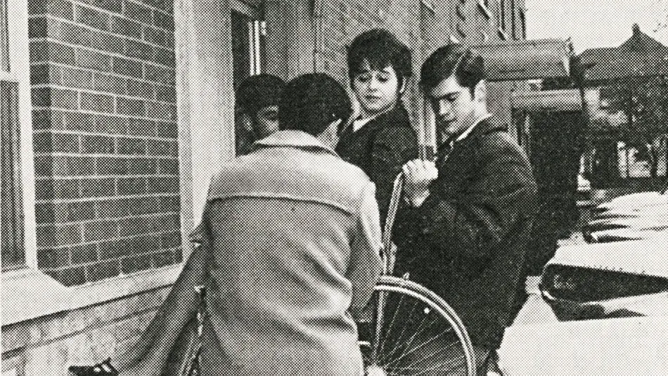 Black and white archival photo of Julie Cochran seated in her wheelchair being lifted by 3 men.
