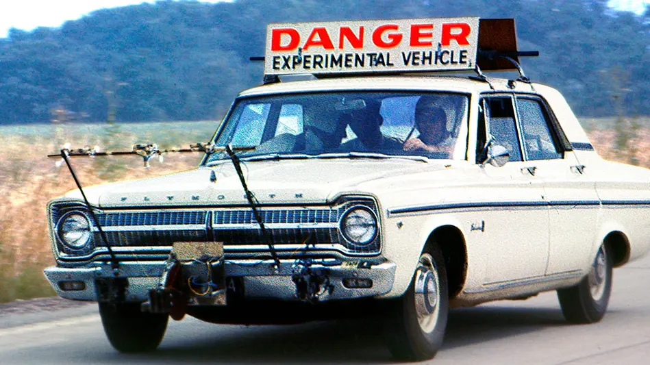 A historic photo of an experimental autonomous vehicle being operated by Professor Robert Fenton. The car is a white 1965 Plymouth sedan with a large sign affixed to the top that reads "Danger Experimental Vehicle". 
