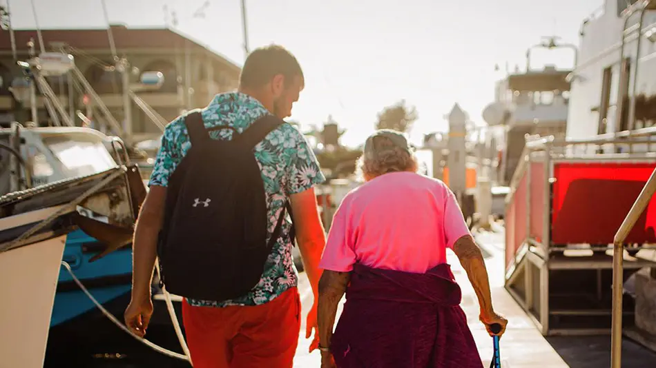 Brad and Grandma Joy are walking away from the camera along a dock surrounded by boats. Brad is wearing a black backpack and Grandma Joy is walking with a cane.
