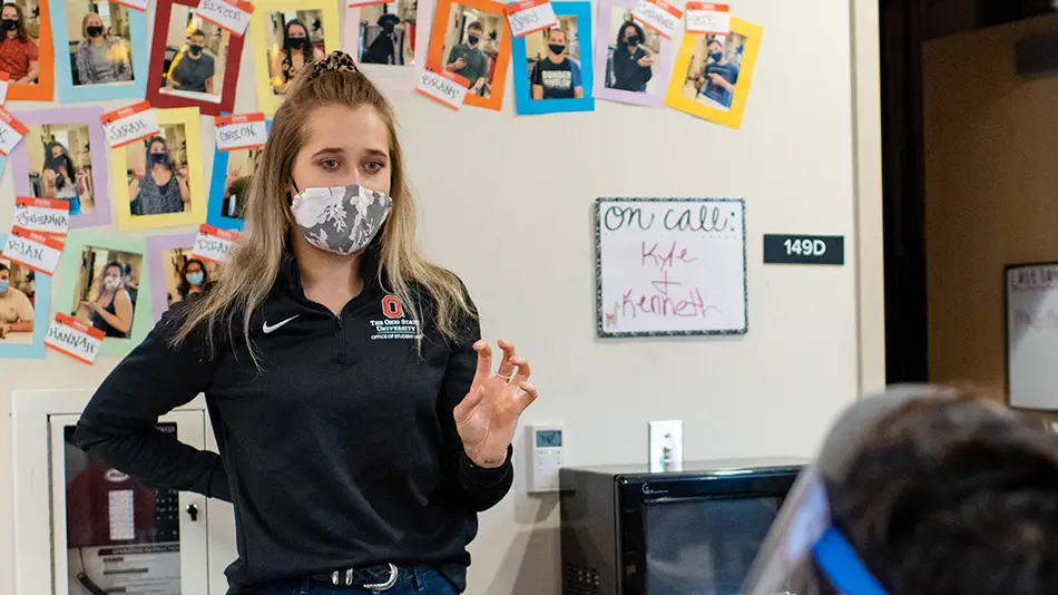 Haley Quinlan is standing with her hand in a claw shape. She is wearing a gray patterned face mask and a black long sleeve polo.