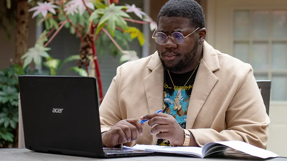 Kenshawn Simmons is sitting outdoors at a table with a laptop and a notebook in front of him. 