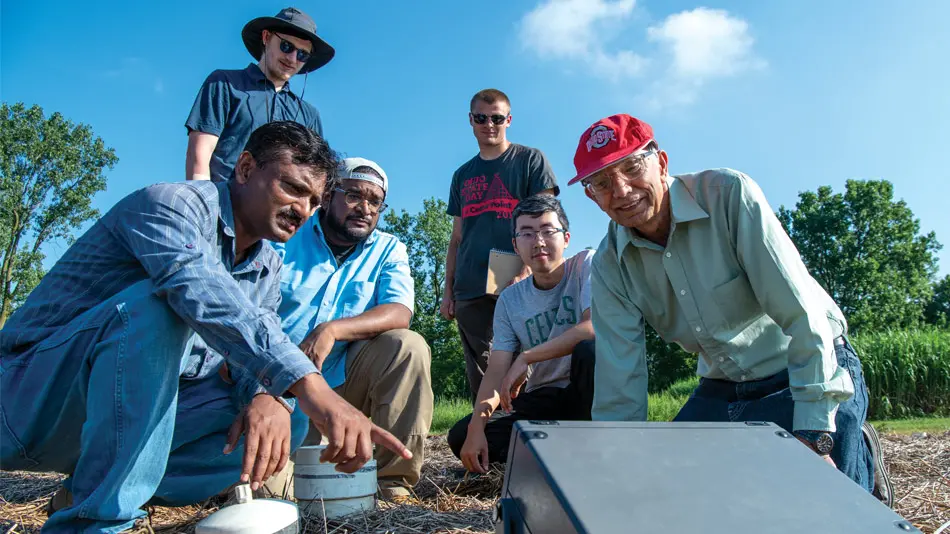 Rattan kneels on the ground in front of a gray electronic box with 5 graduate students and researchers.