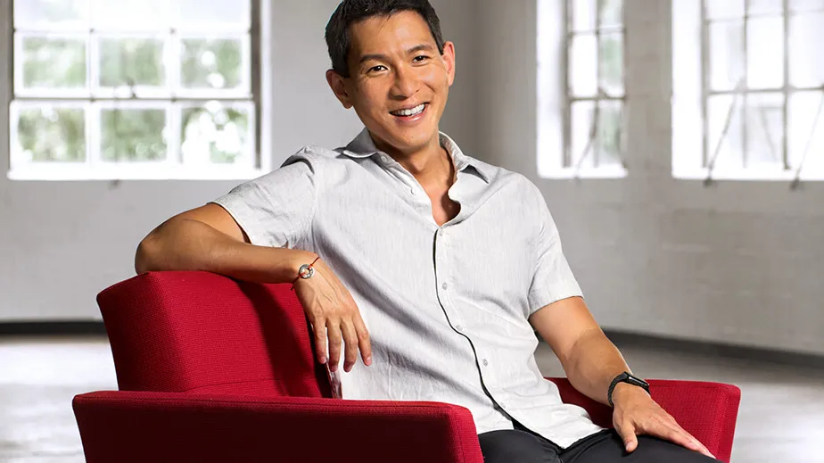AAPI man sitting in red chair wearing short sleeve button up shirt. 