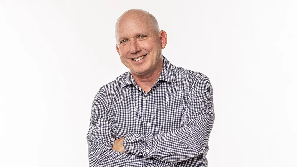 A white man with a shaved head stands confidently with a big smile and arms crossed.