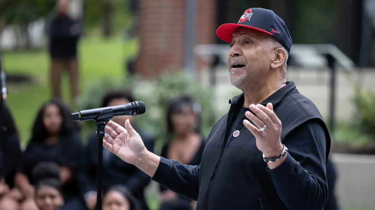 A Black man wearing an OhioState ball cap gestures with hands spread wide as he speaks to an audience--only vaguely suggested in the background--at the dedication ceremony. He looks optimistic and proud. 