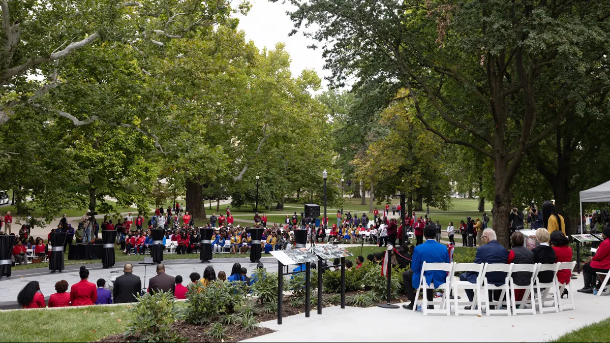 A farther-away view of the plaza dedication shows a sizable crowd gathered around an oval-ish paved plaza. Along the edge stand shoulder-high pillars that each represent a fraternity or sorority. On land higher up than the plaza is a space where those who will speak at the ceremony sit and from where speakers can address people on the plaza.