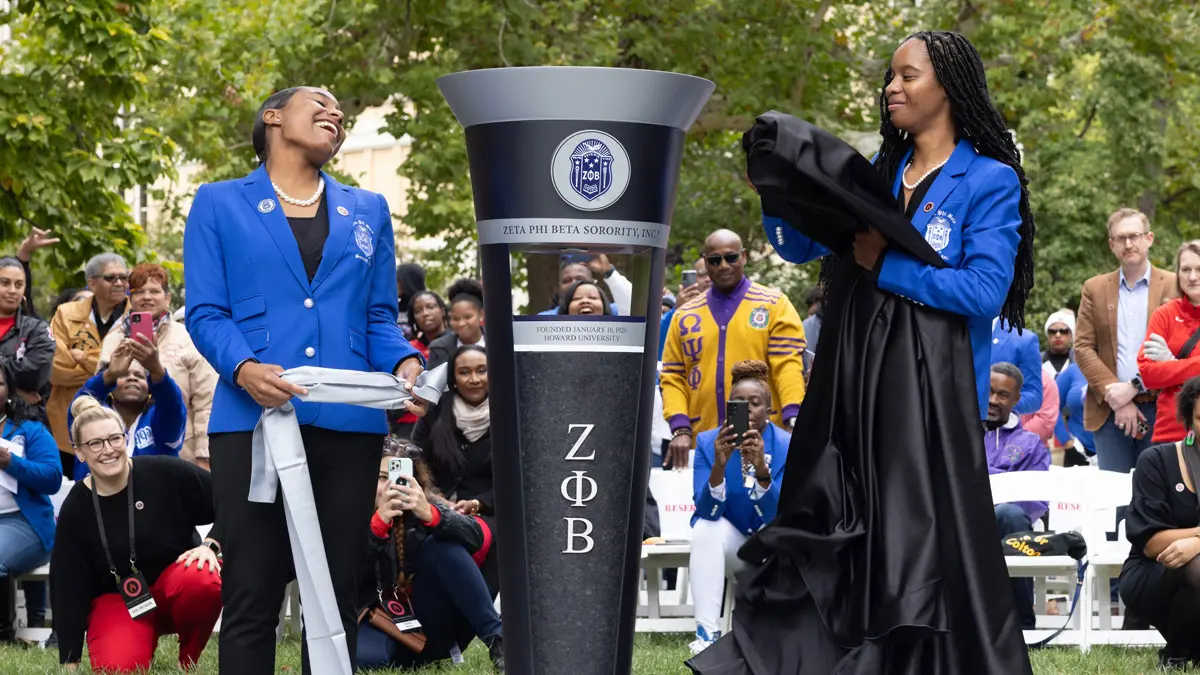 Two young women wearing blue suit jackets unveil their sorority's pillar along the edge of the plaza. One young woman has her head thrown back as she laughs; the other watches her friend happily.
