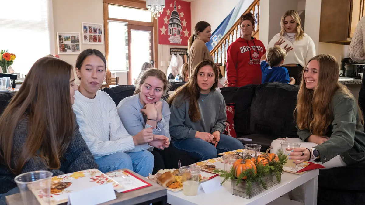 Four young women on a coach and one more in a chair sit around a coffee table. They make eye contact as they listen to one another talk.