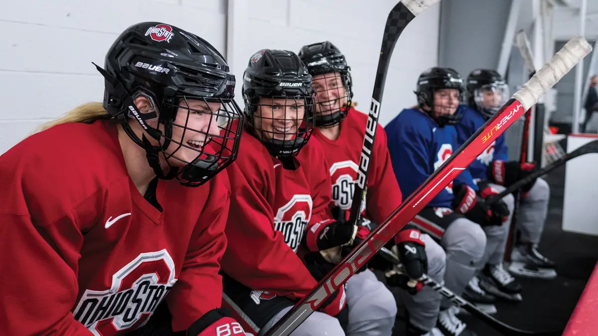 Sitting on a bench during practice, helmeted ice hockey players hold their sticks and laugh and talk while watching the ice (not shown in the photo). The first three wear red jerseys and the last two wear blue, but all have big Ohio State logos on the chest.