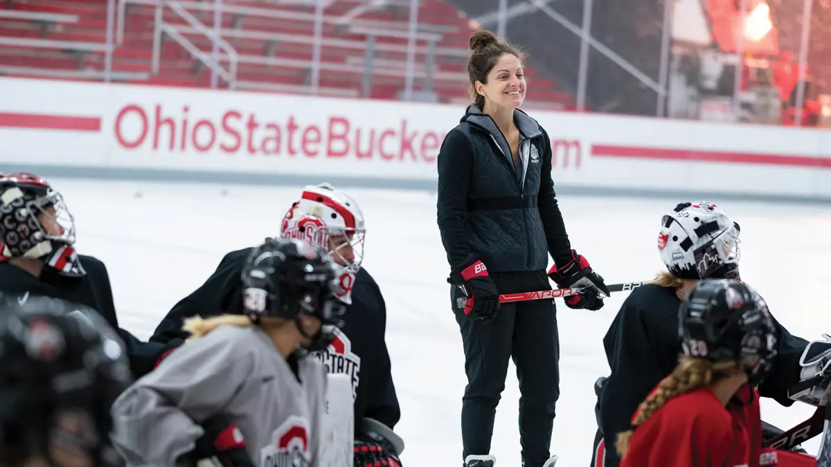 In an ice rink, a woman in black workout clothes and ice skates holds a hockey stick while standing behind crouching hockey players. She’s grinning.