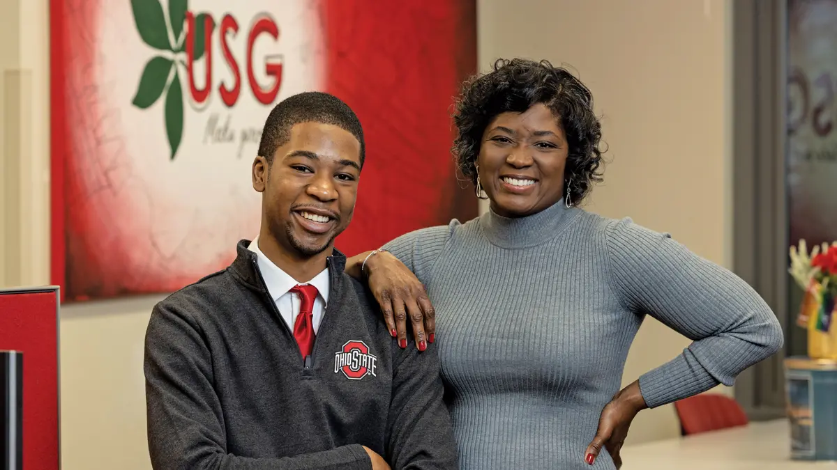 A young black man poses with his mom. He’s wearing a button down, red tie and Ohio State pullover, and she looks stylish in a gray dress. They both wear genuine smiles, as evidenced by the way their eyes crinkle.