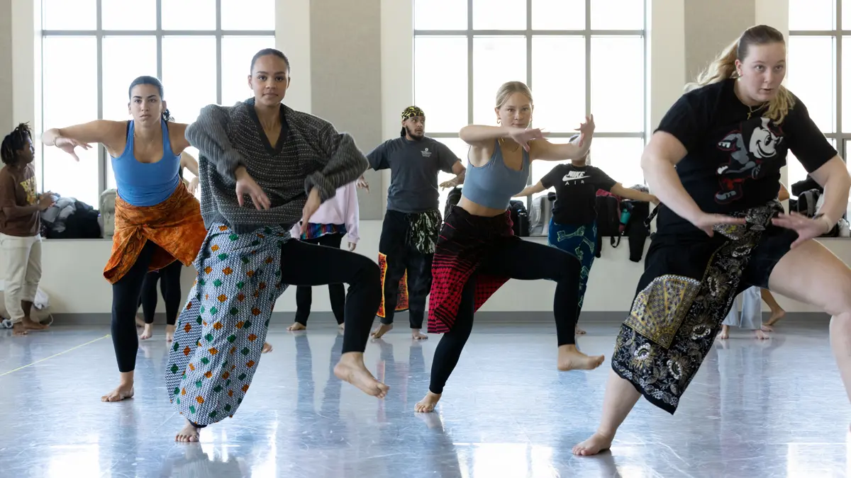A group of young women lift their left legs and bend their arms the way you’d do a pushup as they dance in a class. Behind them are huge windows casting light through the room and a line of younger dancers. Each dancer is adorned with an African patterned fabric tied around their waist.