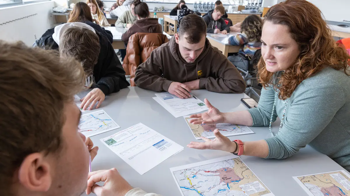 A female teacher with curly hair makes eye contact with a male student as she uses her hands to intently explain a concept. They’re at a table with maps and two ither male students, and behind them, are other tables with students. Bright light from big windows filles the classroom.