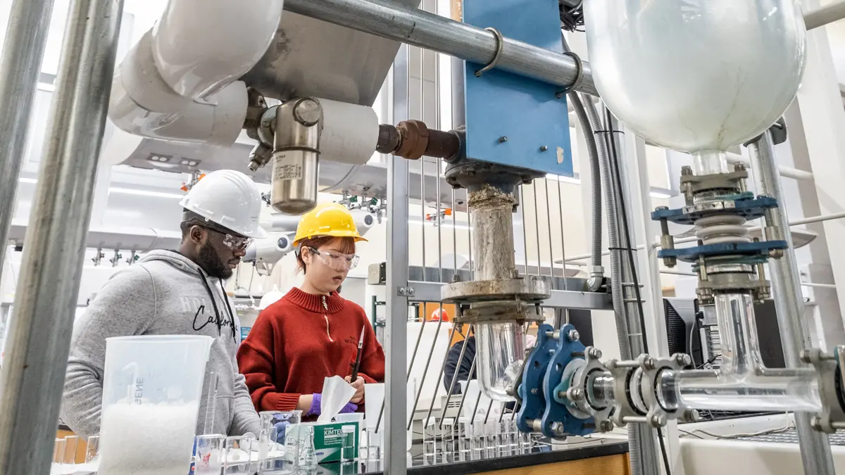 Two students, an undergrad and a teaching assistant, work at a lab table connected to towering metal framework and giant tubes. Machinery and large lab equipment surrounds them. Both wear hard hats and safety glasses.