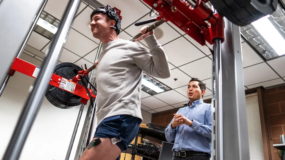 Wearing metal headgear and other body sensors, a young man power lifts a bar with big weights above his head while standing within a metal  structure. He grimaces as he bears the weight. A man behind him in dress clothes looks pleased as monitors his movements and talks.