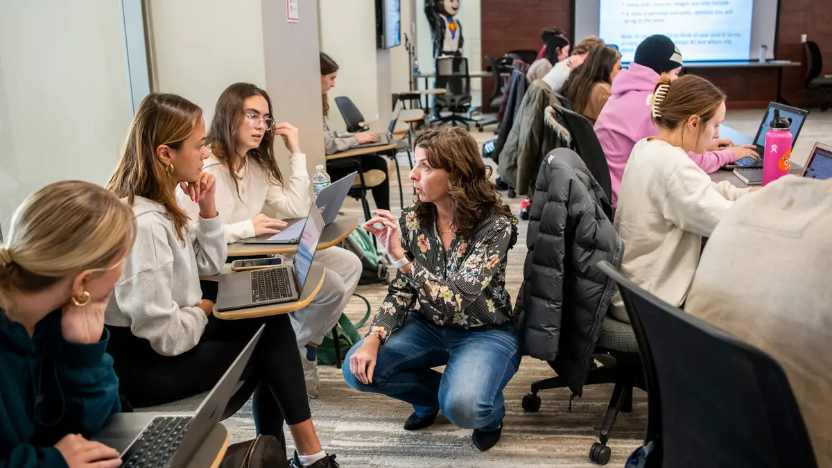 A professor crouches as to get on the same level as students sitting in seats along windows. She’s talking to two females intently listening. Another row of students sits at a long table behind her. Each student has a laptop in front of them and they are all hard at work on an assignment.