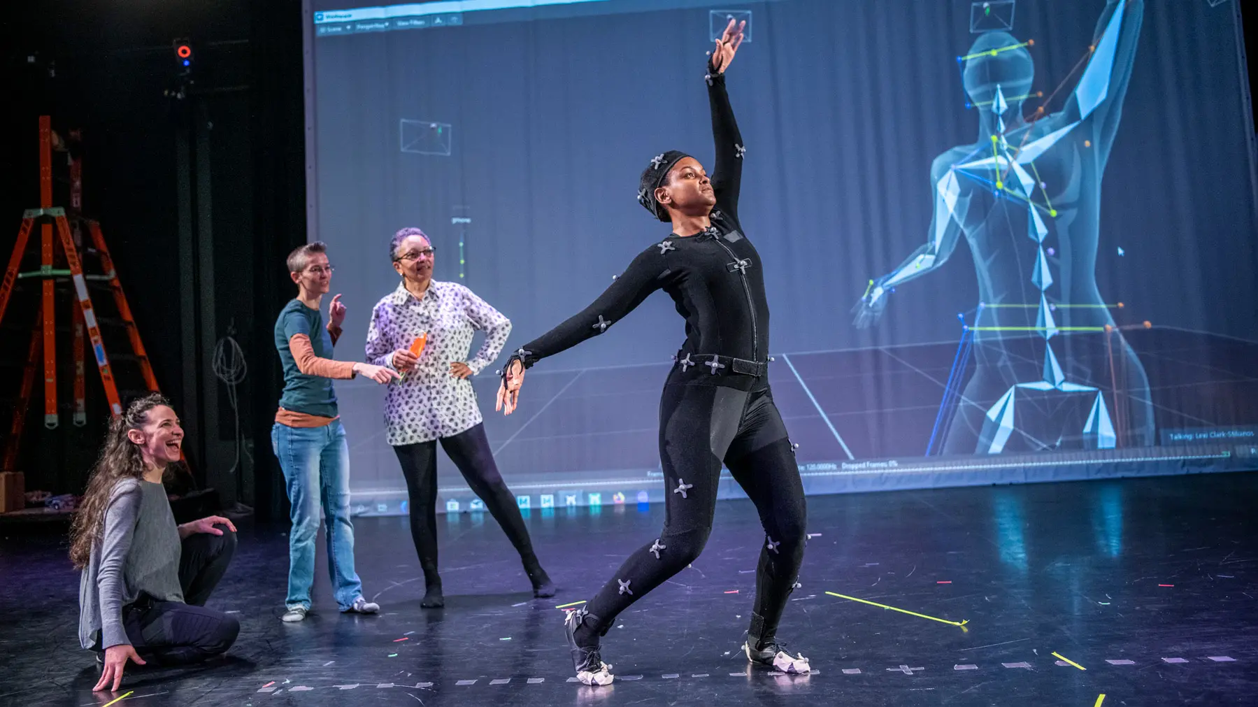 On a stage, a dancer in a black body suit and sensors steps forward and elegantly spreads her arms as three women behind her watch excitedly. A computer-generated geometrical illustration of the dancer highlights where her bones are as she moves.