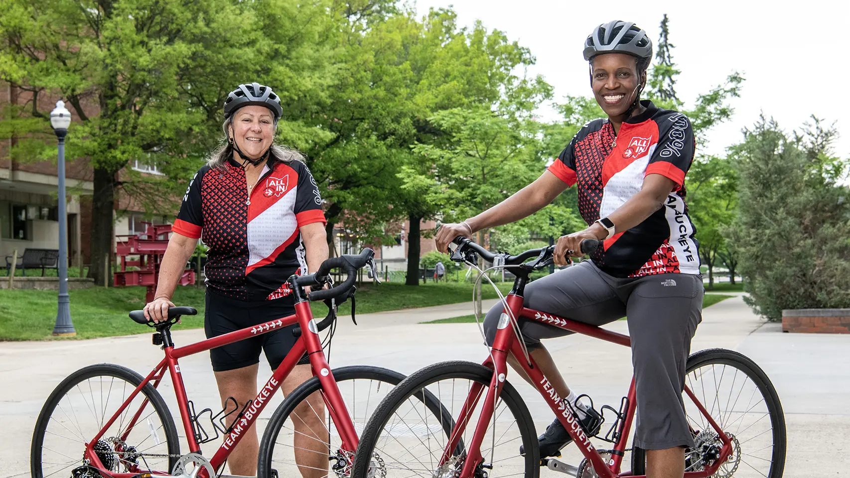 Two smiling women stand with their matching bicycles wearing matching Team Buckeye jerseys and bike helmets. On the left is Molly Ranz Calhoun, a tall white woman, and on the right is Provost Melissa Gilliam, a tall Black woman.