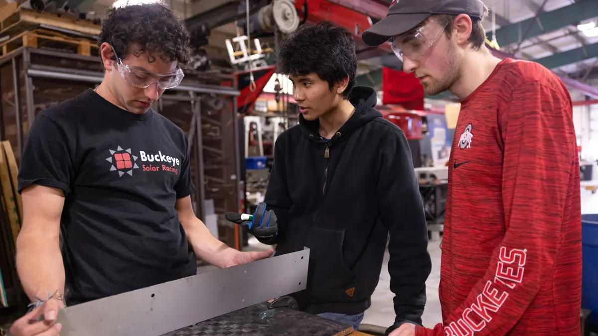 A young man holds a flat, thin piece of metal about 6 inches tall and almost twice as wide as his body as two others look at it or him intently. The middle man gestures as he talks.