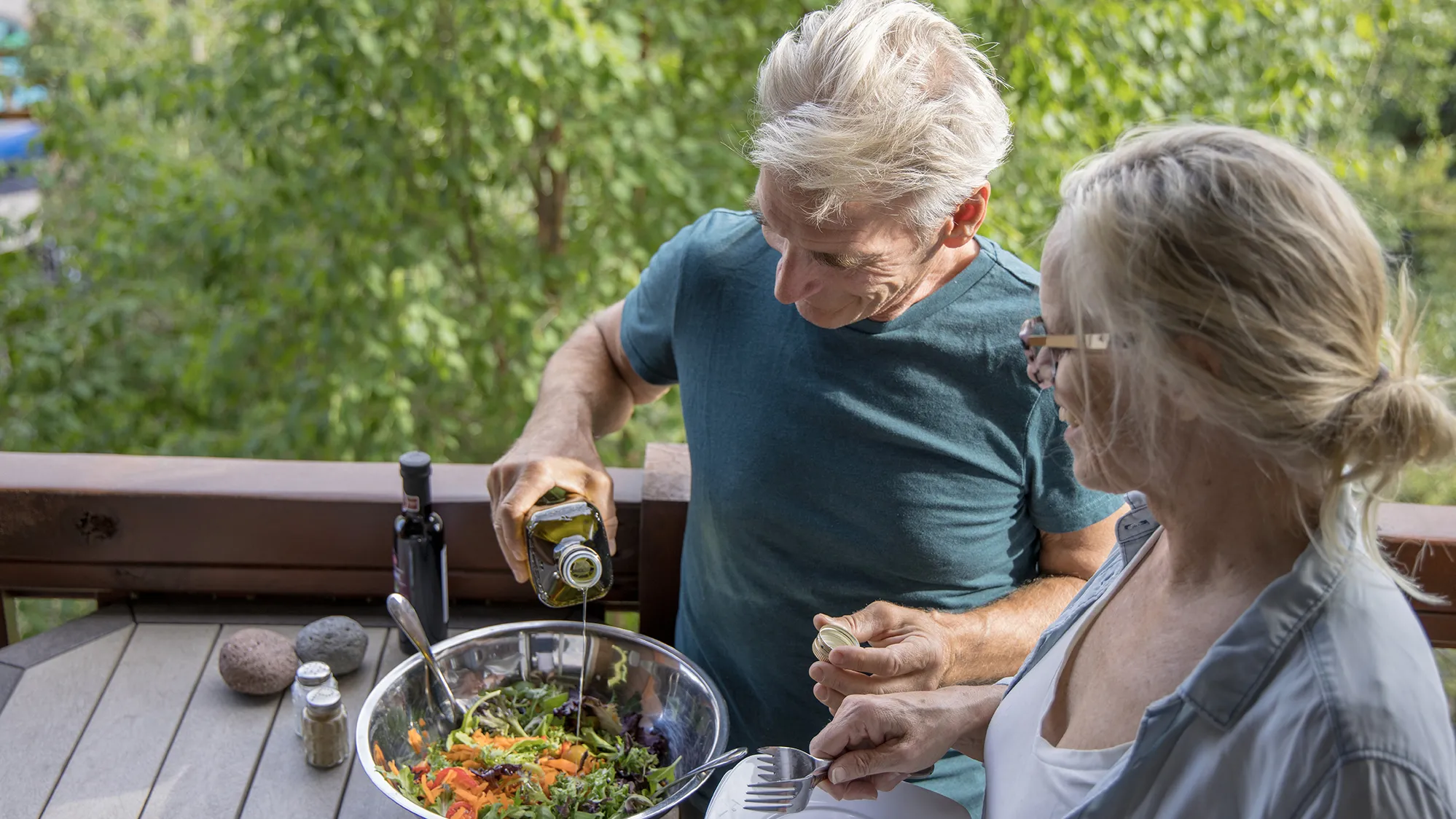 Outside on a porch or deck, two older white people stand around a metal mixing bowl with a salad of greens, carrots and cherry tomatoes. The man is drizzling olive oil onto the salad and the woman holds plates and forks. Because the photo was taken from slightly above them and they’re both looking down at the salad, you can’t see their eyes.