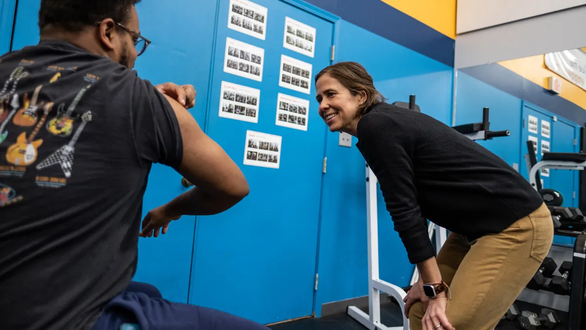 Carmen Swain, a thin white woman, smiles as she leans down, hand on knees, to listen to a young Black man with glasses whom we see from behind. He has the sleeve of his T-shirt pulled up and gestures at his bicep.