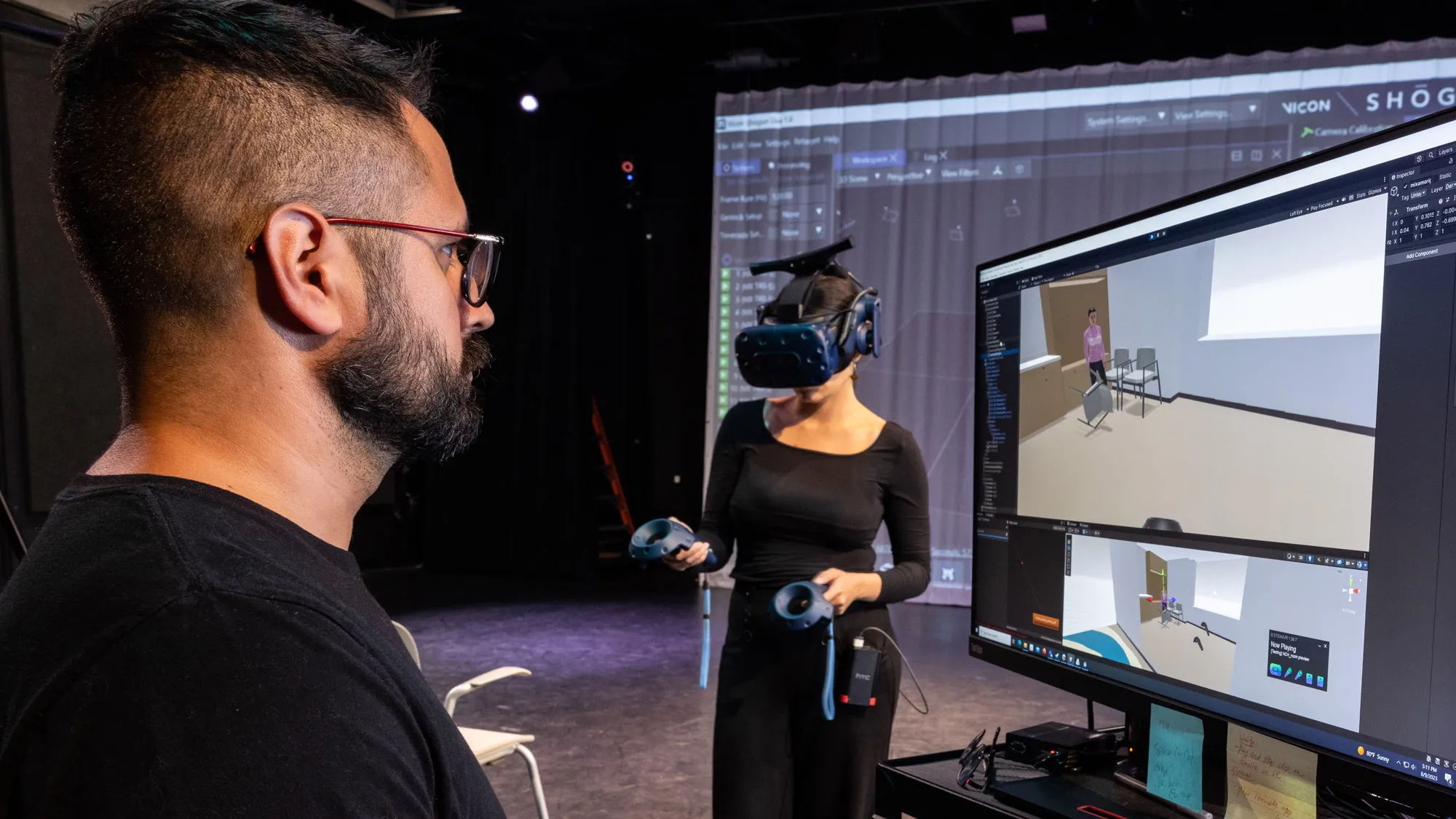 A man works on a computer whose screen shows a classroom setting while a woman on a stage beyond him wears a virtual reality headset and sensors used for motion capture. Ultimately, her movement will be shown on the screen he works on. 