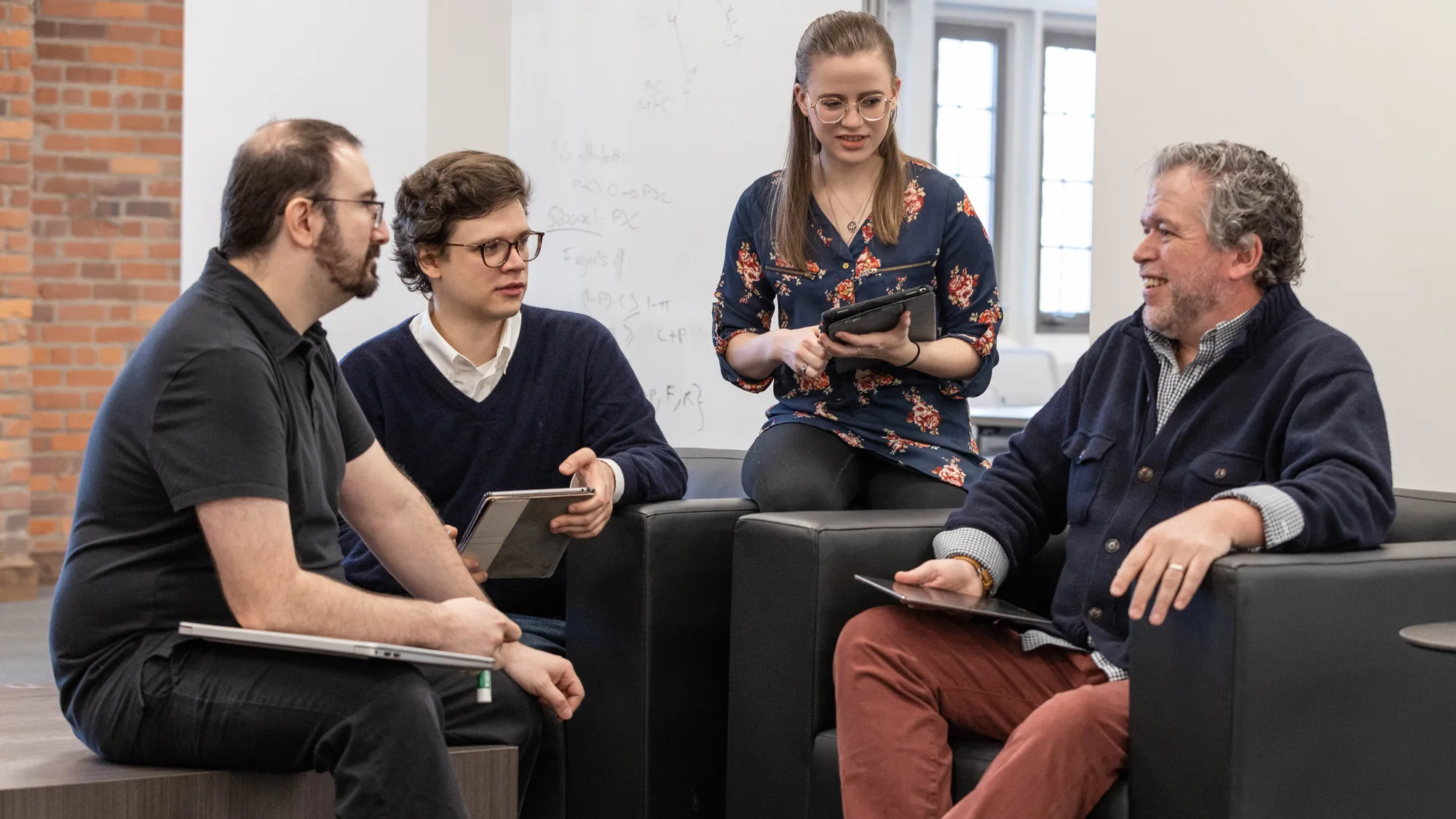 Bear Braumoeller sits in a chair as he speaks with three graduate students sitting around him. He has curling, graying hair and wears a cardigan and loafers, and his skin crinkles around his eyes as he smiles. 