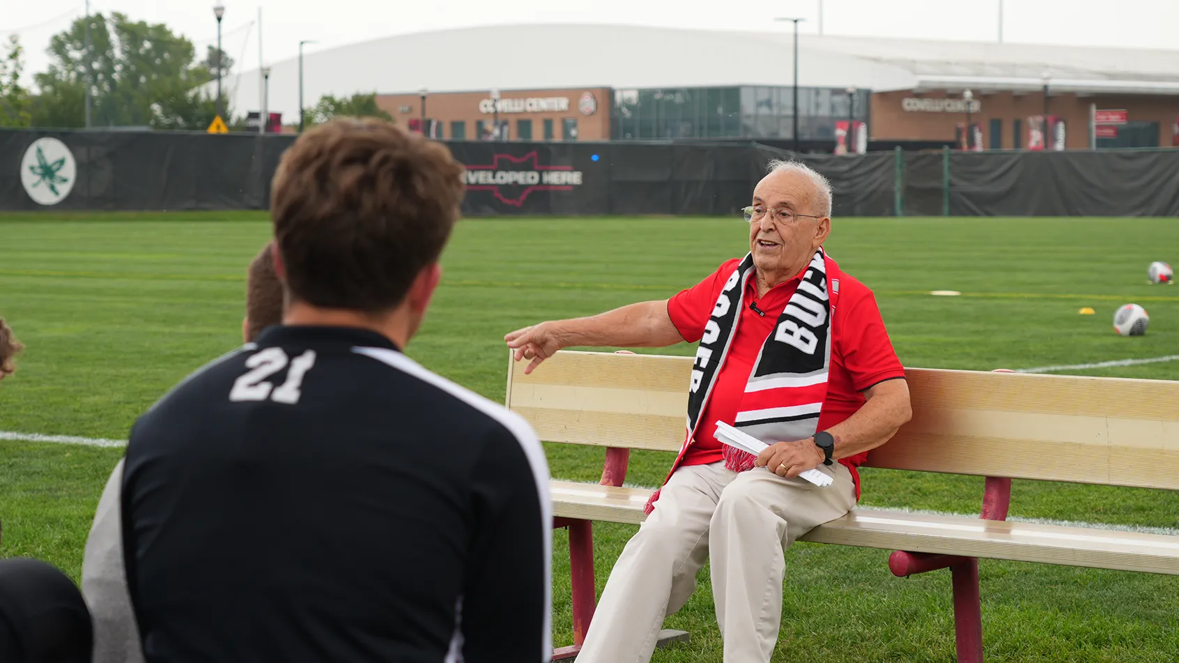 An older man happily chats, while wearing a scarlet shirt and long Buckeyes scarf, lounging on a bench along a soccer field at the Schumaker Complex. Behind him, the grass looks vibrant and soccer balls and field markings can be seen. Facing him are members of the men’s soccer team listening as he talks.