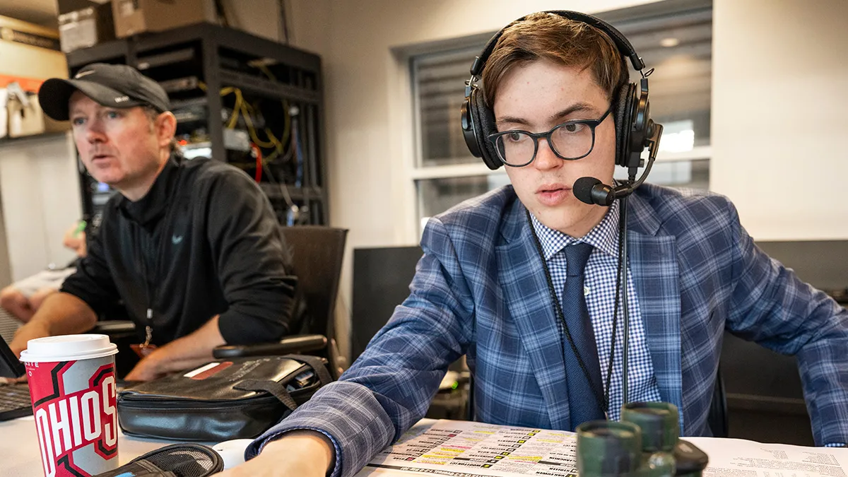Wearing headphones with a mic, a checked sport coat and tie, Tyler Danburg focuses on game action off screen as he broadcasts a game from a the press box in a stadium.