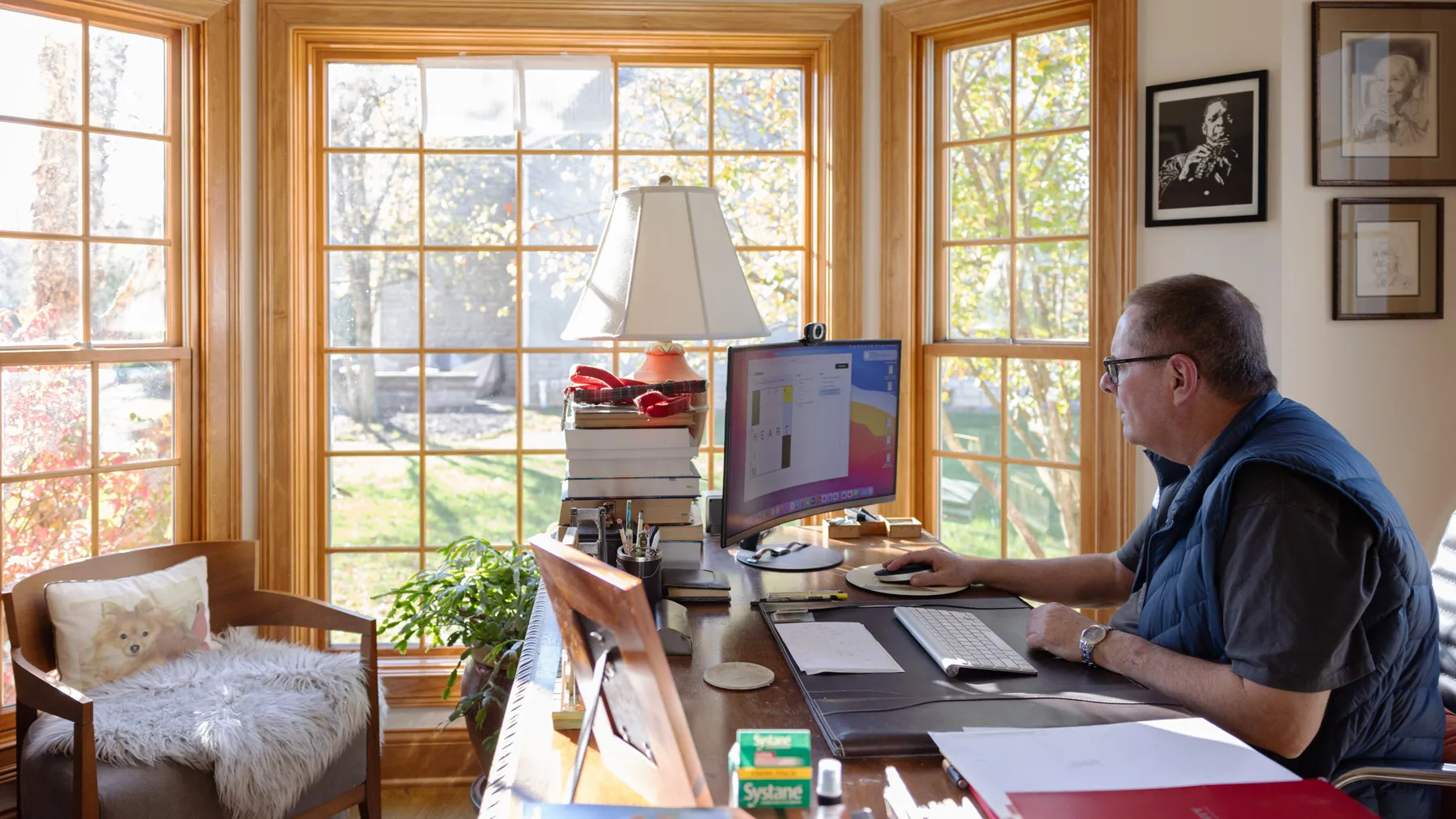 Ohio State Professor Lou DiMauro completes an online crossword puzzle in his home office. Hanging on the walls are jazz and sports photos and a nearby chair has a pillow with a picture of a dog on it. Lou focuses on his screen, leaning slightly toward it.