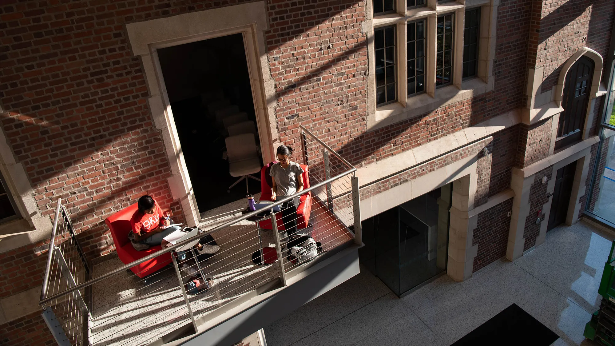 This photo shows Pomerene Hall on Ohio State’s campus. Inside, the lights are all on, so the windows glow—a geometric-pattern mural can be seen inside a wall of ground-to-roof windows. Students wearing backpacks pass by on the sidewalk in front.
