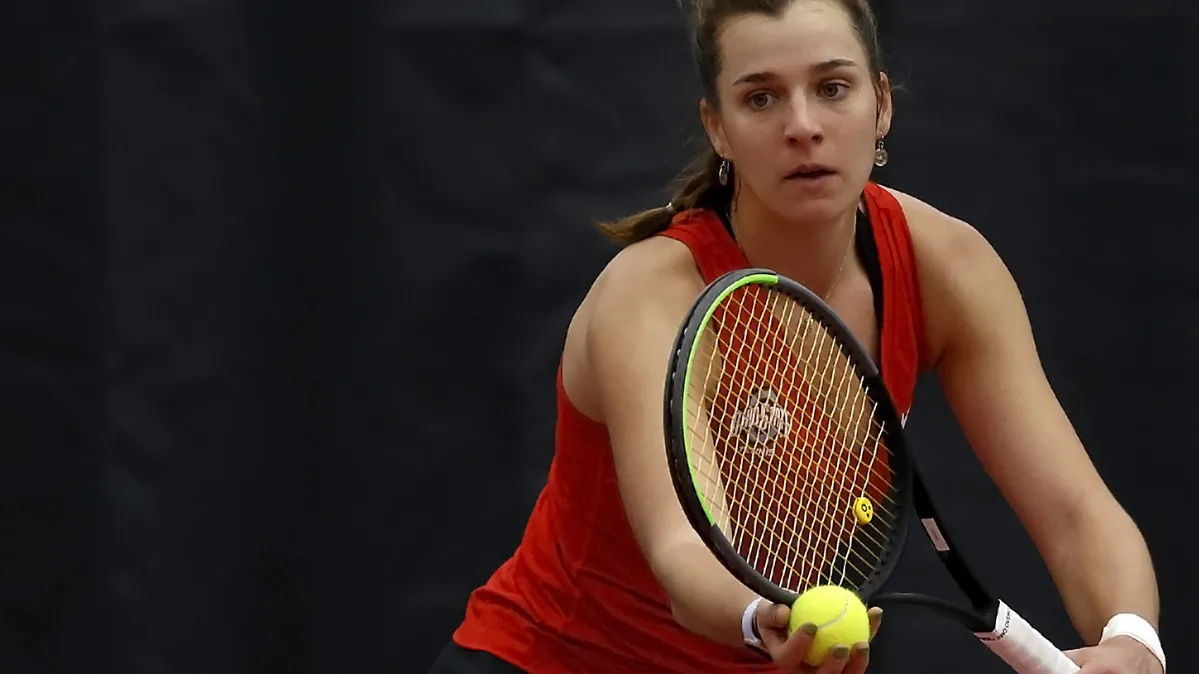 On the court, Irina Cantos Siemers, an Ohio State tennis player, holds a ball in her right hand and her racket in her left, getting ready to serve. She seems focused on her opponent’s side of the court as she prepares for action. It’s a sunny day, so she wears a visor and sleeveless uniform.