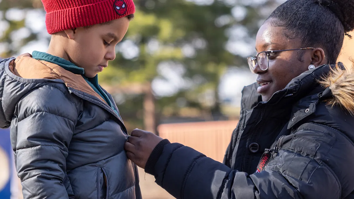 On an outdoor playground, a college student smiles as she crouches down to zip a child’s coat. She’s also wearing a heavy winter coat (along with glasses and her hair piled in a big bun on top of her head). The boy, wearing a stocking hat and gloves, watches her hands.
