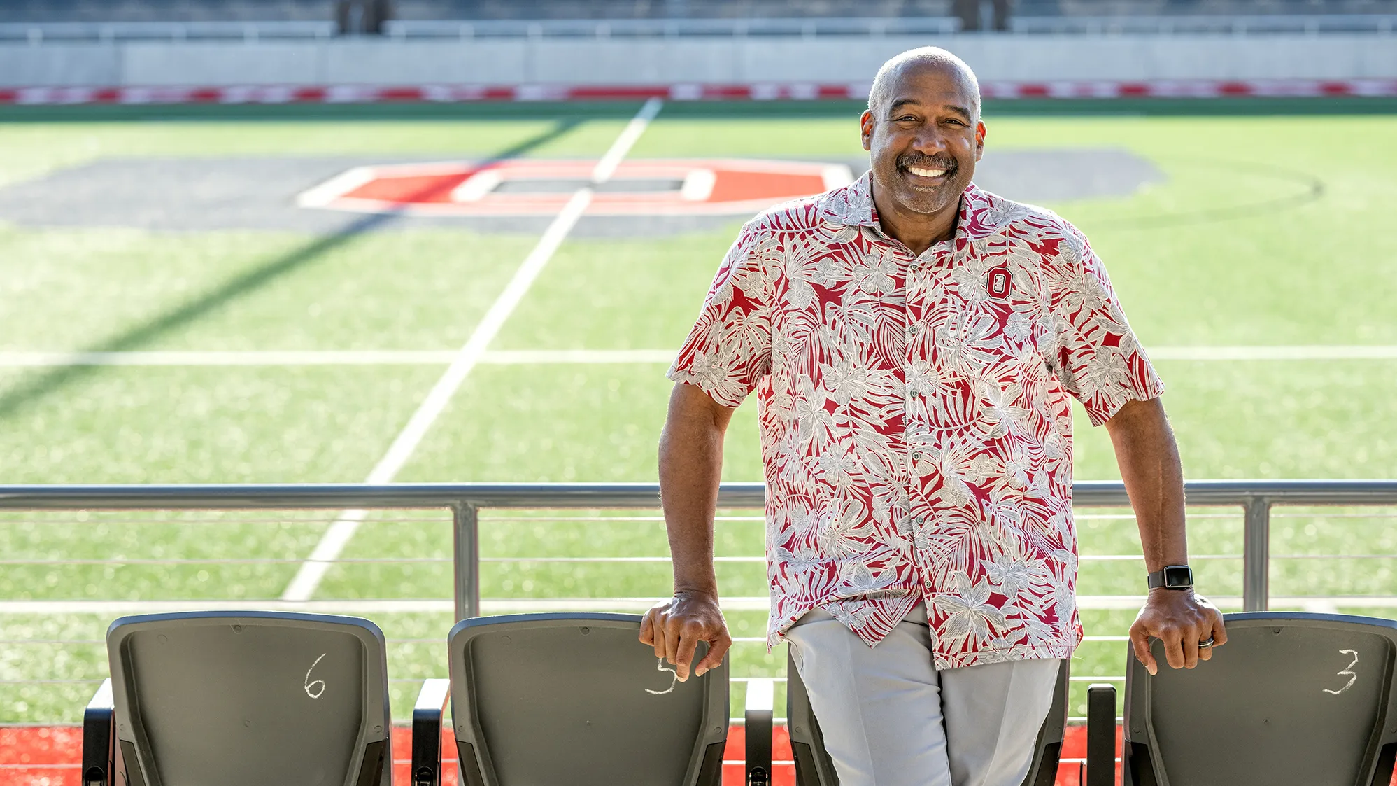 Wearing an Ohio State-colored tropical shirt—that looks perfect for retirement or vacation—Gene Smith grins as he leans on some front-row seats at Ohio Stadium. 